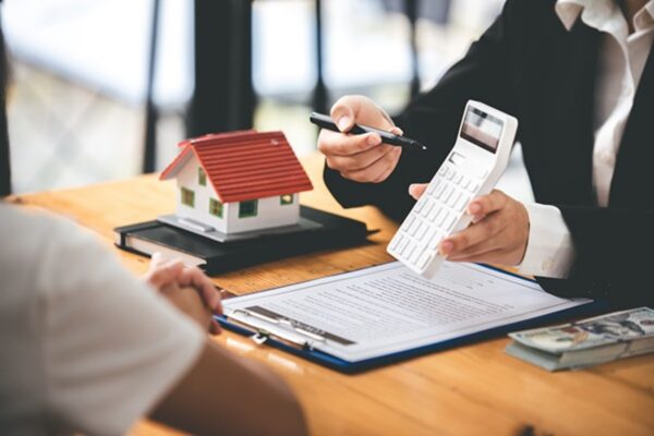 A real estate professional in a black blazer holding a white calculator and a pen over a contract, with a small model house and a stack of cash on the wooden desk.